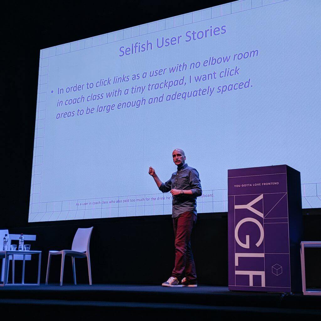 Adrian Roselli on-stage at YGLF Lithuania. Slides read “Selfish User Stories: In order to click links as a user with no elbow room in coach class with a tiny trackpad, I want click areas to be large enough and adequately spaced.”