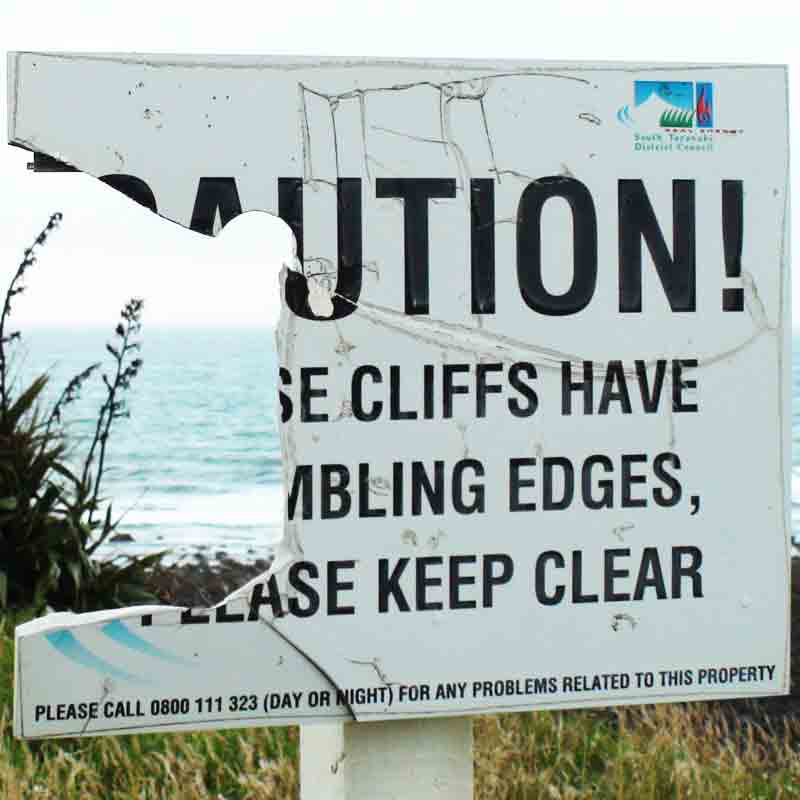 A sign near a cliff, broken nearly in half with the left side missing. The visible text is “UTION! e cliffs have mbling edges ase keep clear.”